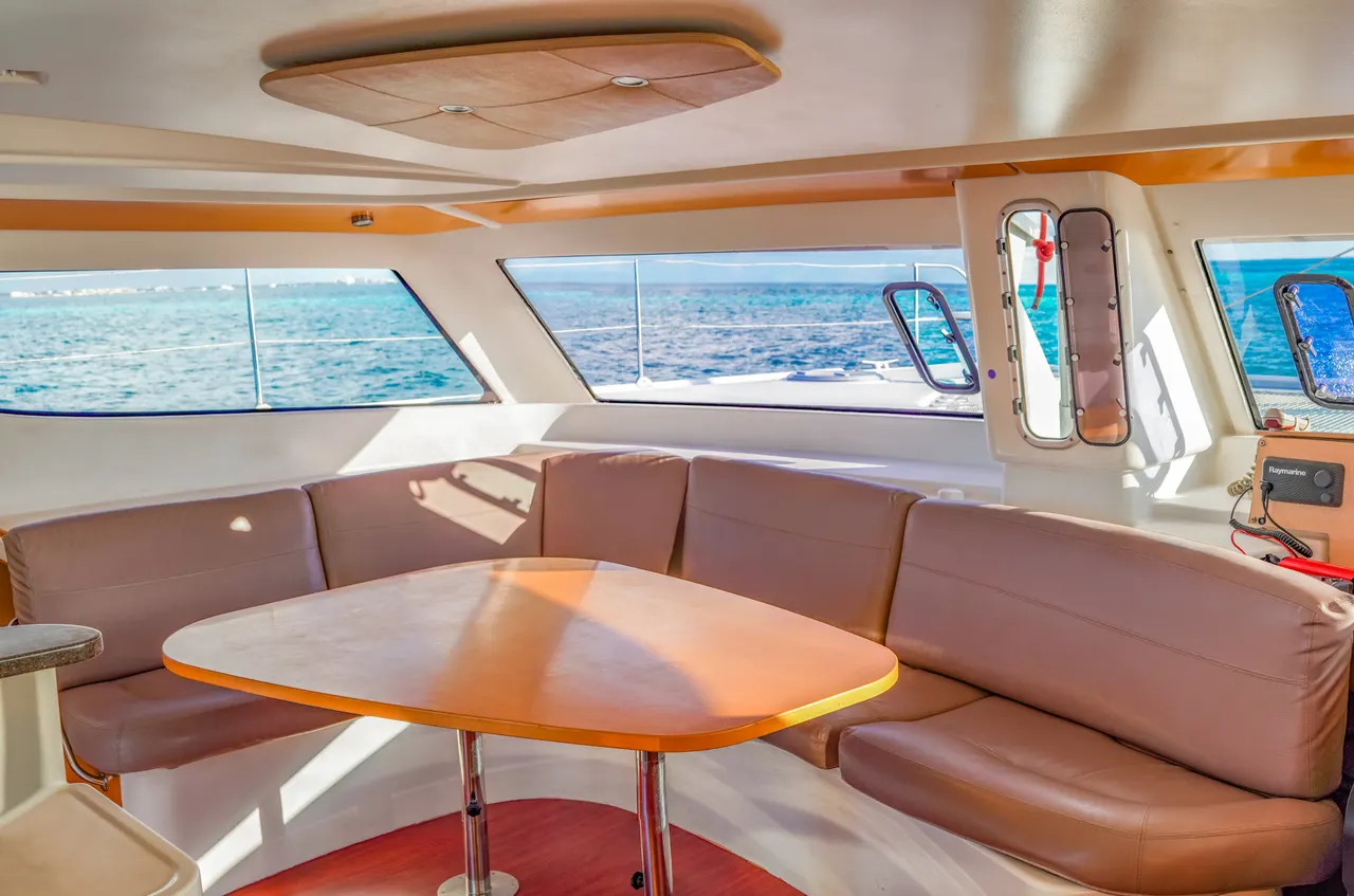 Interior luminoso de un catamarán de lujo con salón equipado con cómodos asientos de cuero marrón, mesa de madera con detalles naranjas, y amplios ventanales que ofrecen vistas panorámicas al mar azul Caribe. Luz natural abundante destaca la elegancia y comodidad del espacio interior.